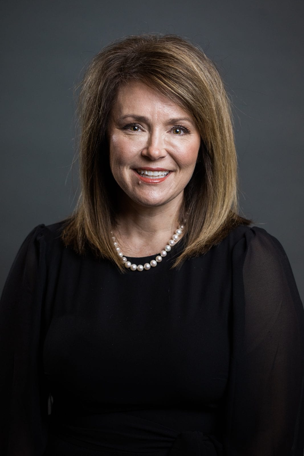 Professional headshots Houston: woman in a black top and pearls on a gray backdrop