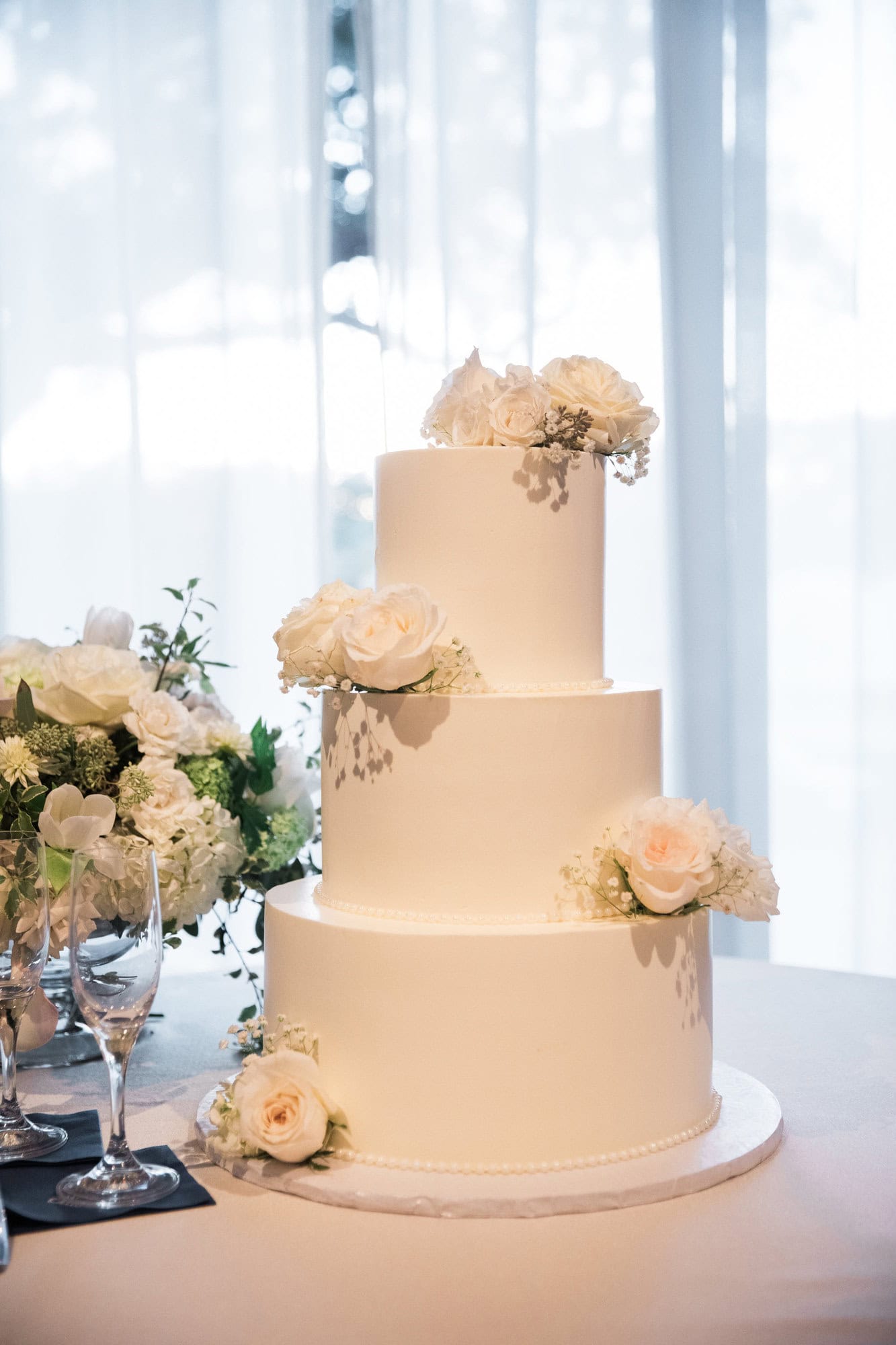 Three-tier white wedding cake decorated with white flowers beside a window.