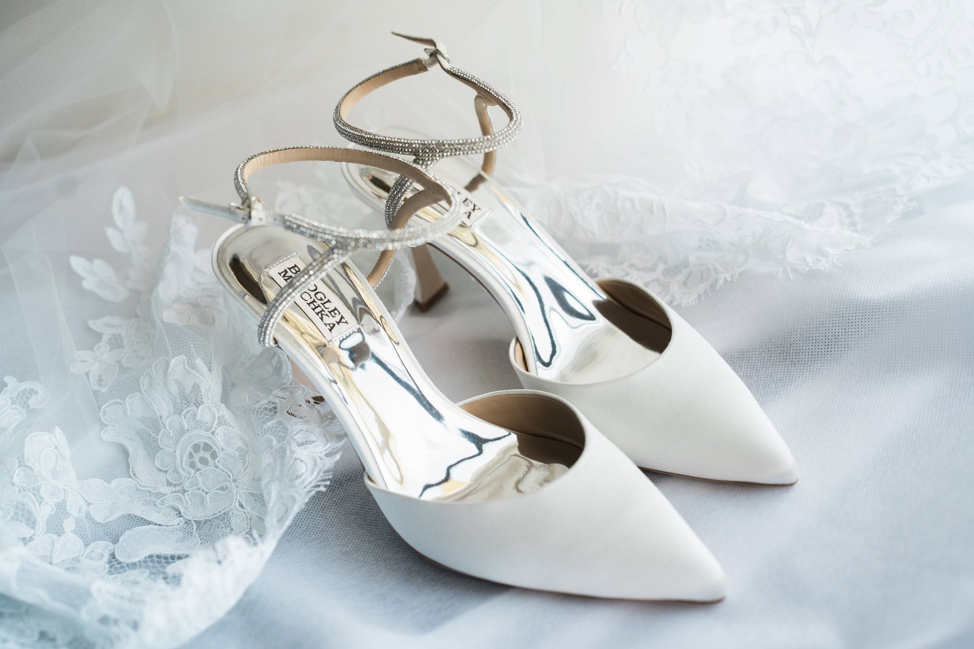 White pointed-toe wedding shoes with sparkling ankle straps on lace fabric.